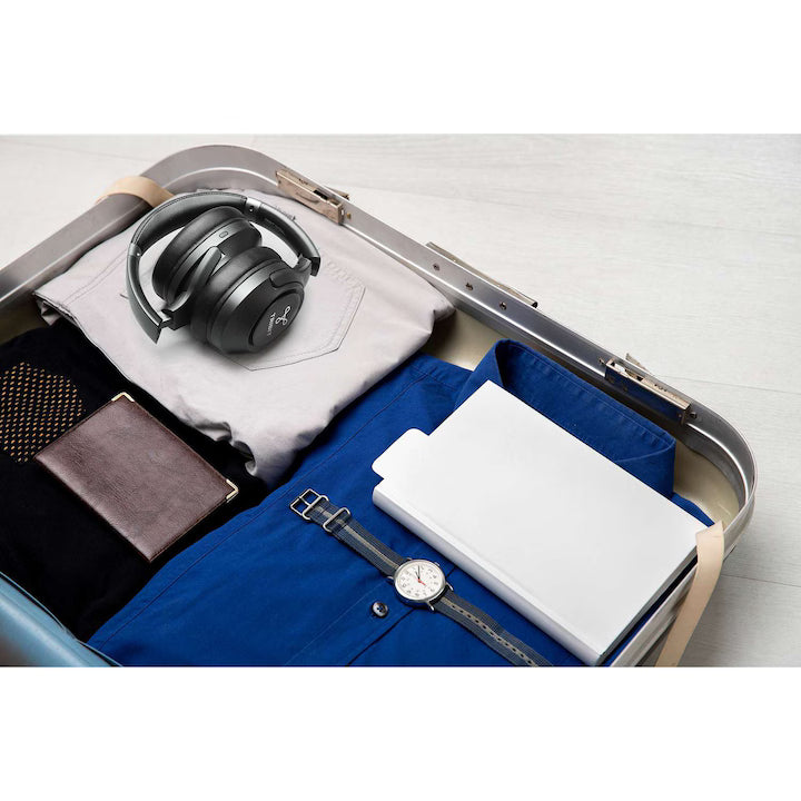 Valiză de călătorie deschisă pe o podea de lemn, conținând căști wireless (model Tribit XFree Go S), un portofel din piele și un ceas. Ideal pentru navetă și călătorii.
